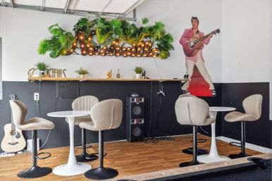 Cozy Nashville-themed music café interior with round white tables, beige swivel chairs, acoustic guitar, speaker stack, illuminated marquee “Nashville” sign with tropical greenery, and a guitar-playing singer mural.