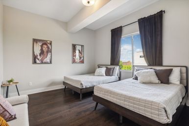 Cozy modern twin bedroom in an urban apartment with two beds in gray striped bedding, fluffy throw pillows, dark hardwood floors, framed wall art, and a window with dark curtains overlooking blue-sky city views.