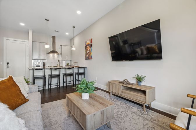 Bright modern open-plan living room and kitchen with wall-mounted flat-screen TV above a wooden media console, wood coffee table with potted fern, gray sofa with rust accent pillows, breakfast bar with four black stools and pendant lights, stainless-steel range hood and dark hardwood floors