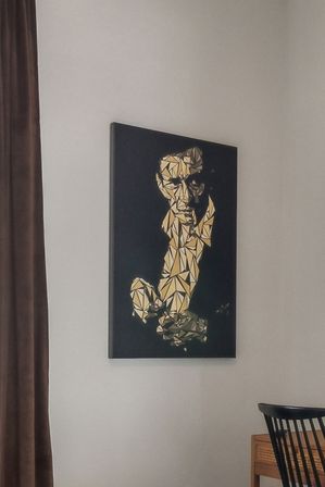 Black-and-gold geometric abstract portrait canvas on a light-gray wall in a modern living room, with a brown curtain and wooden chair visible.