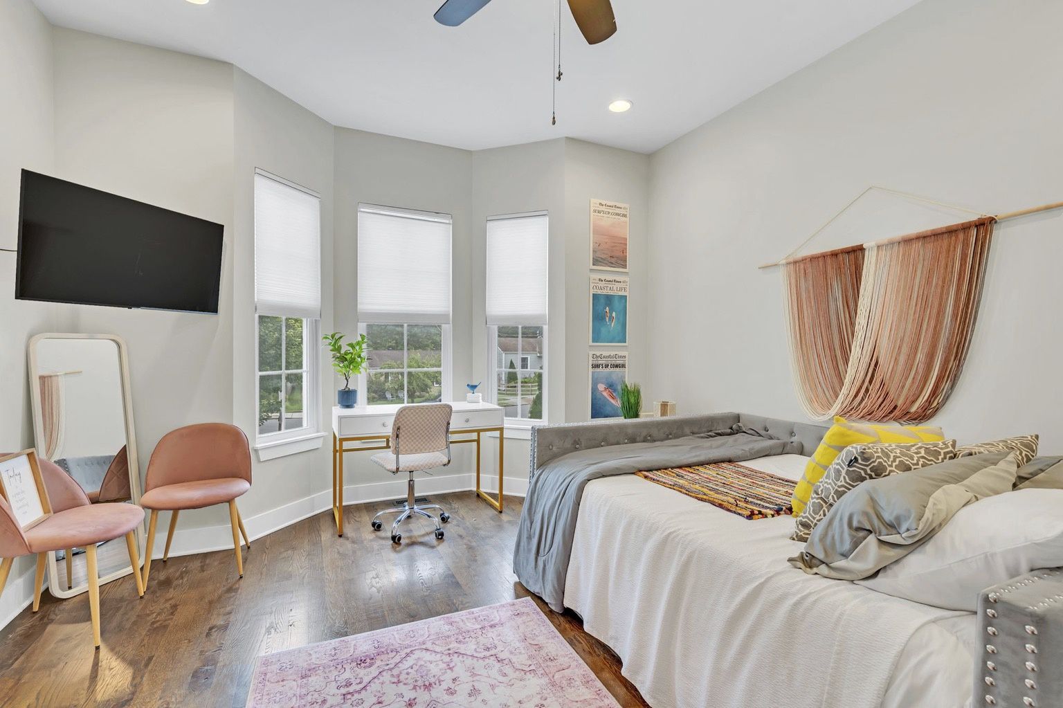 Bright modern bedroom with bay windows and a white desk workspace, swivel chair, wall-mounted TV, blush pink accent chairs and full-length mirror, tufted gray daybed with colorful throw pillows and woven wall-hanging on hardwood floors.