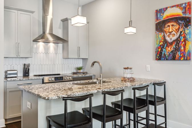 Sleek modern kitchen with speckled granite island breakfast bar, four black stools, stainless steel range hood and stove, pendant lights, and colorful portrait wall art.