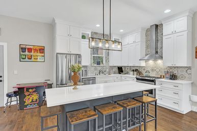 Bright modern white kitchen with large quartz island, five wooden bar stools, brass pendant lights, stainless steel appliances, patterned tile backsplash, hardwood floors and a colorful arcade table in the corner.