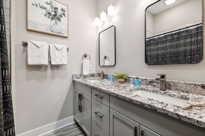 Contemporary residential bathroom with double vanity and marble-look granite countertop, gray cabinets, two rectangular mirrors, brushed metal faucets, white folded towels, patterned black shower curtain, small potted plant and toiletries.