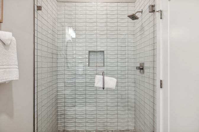 Glass-enclosed modern bathroom shower with white textured 3D wall tiles, square rain showerhead, recessed niche and towel on the glass handle — bright, spa-like residential shower.