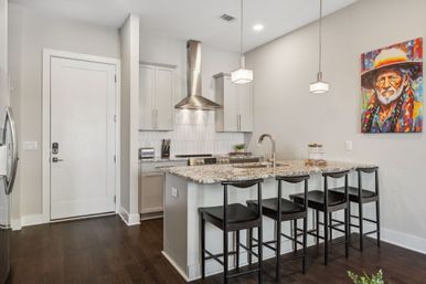 Contemporary open kitchen with granite island breakfast bar, four black bar stools, stainless steel range hood, white cabinets, pendant lights, dark hardwood floors, and colorful portrait wall art.