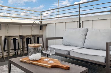 Rooftop patio lounge with light-gray cushioned sofa, metal bar stools, coffee table holding a glass jar of ice and two coupe glasses on a wooden board, with blue sky and railing above.