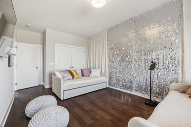 Bright contemporary apartment living room with dark hardwood floors, cream daybeds topped with gold, purple and silver pillows, two knitted poufs and a sparkling silver sequin feature wall.