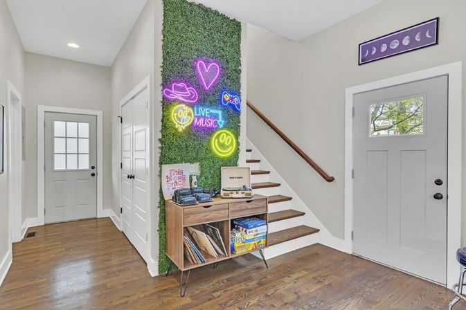 Modern home foyer with wooden staircase, faux moss accent wall and colorful neon signs, mid-century console holding vinyl records and board games on hardwood floors.