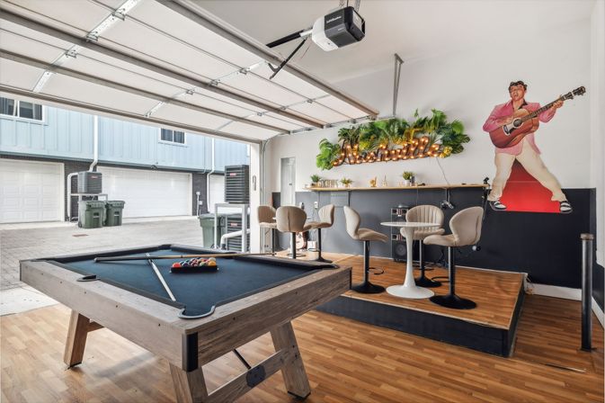Open garage game room with a black-felt pool table in the foreground, raised bar area with round tables and stools, neon "Nashville" sign framed by greenery, and a colorful guitarist mural on the wall.