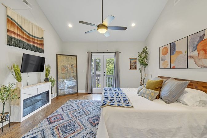 Bright contemporary master bedroom with vaulted ceiling and fan, king bed with decorative pillows and throw, colorful geometric area rug, wall art and woven tapestry, TV on white media console, glass doors to balcony and hardwood floors