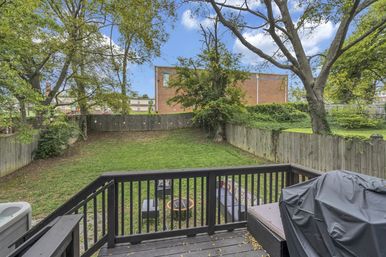 View from a dark wooden deck over a fenced suburban backyard with a covered grill, outdoor seating around a fire pit, grassy lawn and mature trees with a brick building visible beyond.