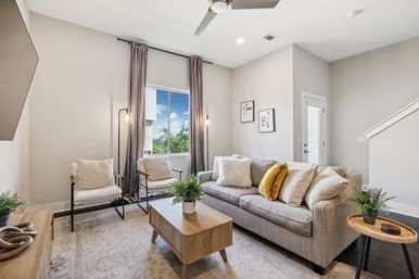 Bright contemporary living room in a suburban home — gray sofa with mustard pillows, wood coffee table, two accent chairs, large window with curtains and ceiling fan.