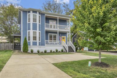 Two-story blue suburban house with bay windows, stacked front porches, a yellow front door and American flag, concrete driveway and a young tree in the front yard.