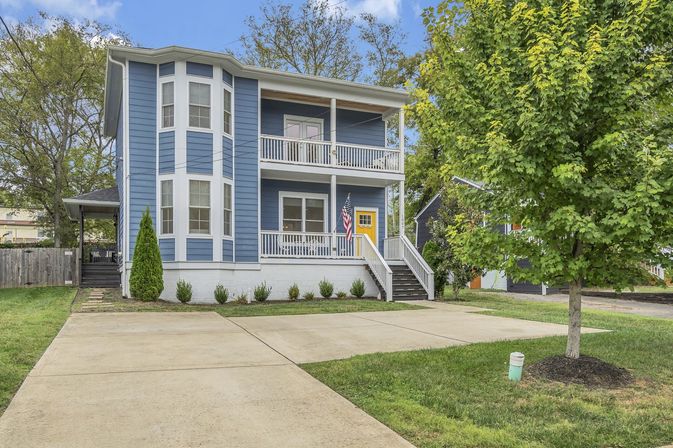 Two-story blue suburban house with bay windows, stacked front porches, a yellow front door and American flag, concrete driveway and a young tree in the front yard.