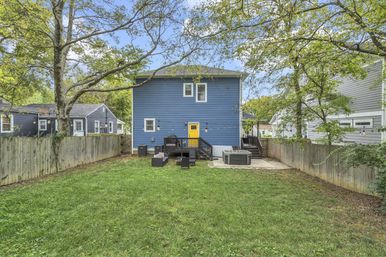 Suburban backyard with grassy lawn and wooden privacy fence, blue two-story house with a bright yellow back door, small black deck with outdoor seating, string lights, and a patio hot tub beneath mature trees.