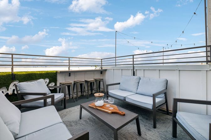 Urban rooftop patio with modern metal-framed sofas and light-blue cushions, low coffee table with ice bucket and glasses, bar stools along railing and string lights under a blue sky