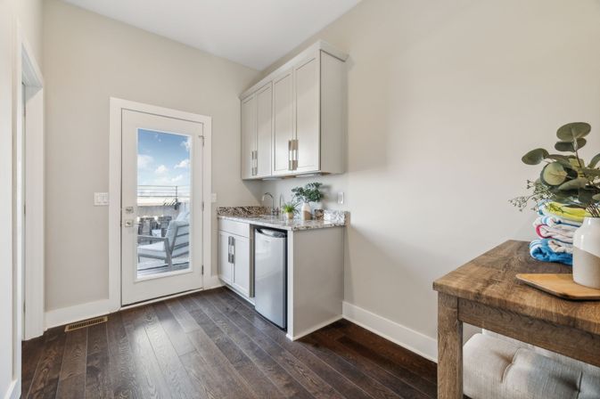 Bright modern mudroom with white cabinets, granite countertop, mini fridge and sink, dark hardwood floors, glass door to a sunny patio, and a wooden table topped with folded towels and eucalyptus