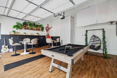 Bright converted garage game room with wood-framed pool table (black felt), raised bar seating and bistro tables, hanging egg chair, acoustic guitar and leafy marquee lights.