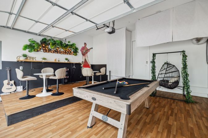 Bright converted garage game room with wood-framed pool table (black felt), raised bar seating and bistro tables, hanging egg chair, acoustic guitar and leafy marquee lights.