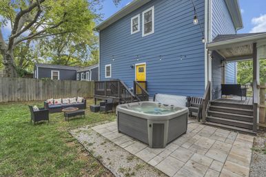Cozy suburban backyard patio with a gray hot tub on stone pavers, blue two‑story house with a yellow door, wooden deck, string lights, outdoor sofa and fire pit, lawn and trees.