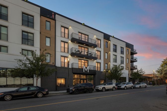 Modern urban apartment building with stacked balconies and street parking at dusk, warm interior lights glowing beneath a pink-blue sunset sky.