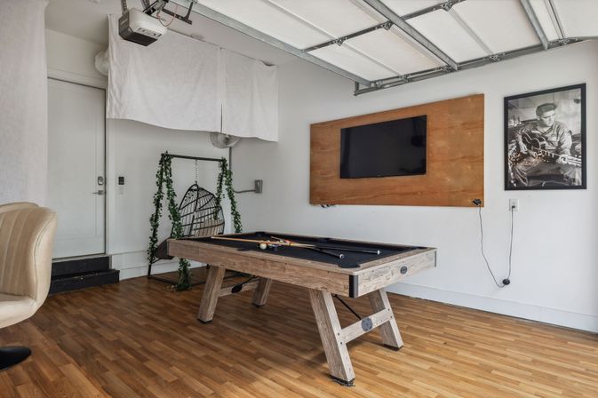 Modern garage game room with rustic wooden pool table, wall-mounted TV on a wood panel, hardwood floors, hanging rattan swing chair draped in greenery, and a framed black-and-white musician print.
