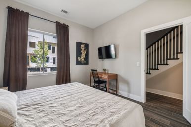 Modern, sunlit bedroom with quilted bed, brown curtains, wall-mounted TV, wooden desk and chair, hardwood floors and visible staircase.