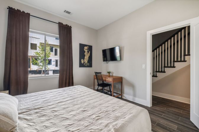 Modern, sunlit bedroom with quilted bed, brown curtains, wall-mounted TV, wooden desk and chair, hardwood floors and visible staircase.