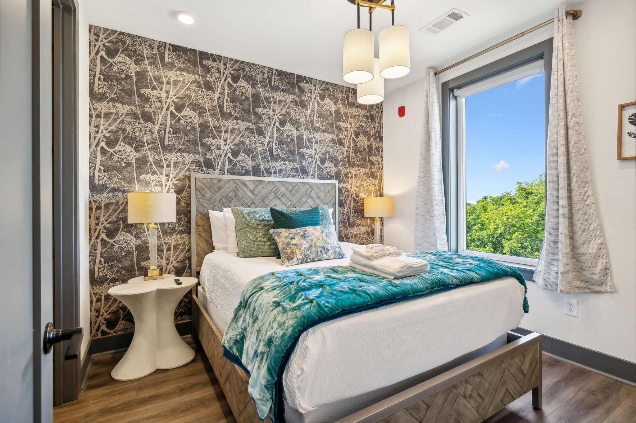 Cozy modern bedroom interior with wood-patterned headboard, botanical wallpaper accent wall, teal velvet throw and decorative pillows on a white bed, bedside lamps and a large window showing green trees and blue sky.