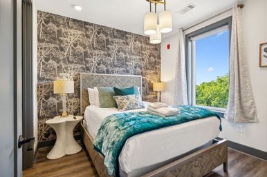 Cozy modern bedroom interior with wood-patterned headboard, botanical wallpaper accent wall, teal velvet throw and decorative pillows on a white bed, bedside lamps and a large window showing green trees and blue sky.