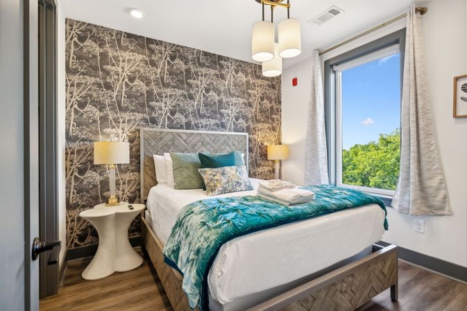 Cozy modern bedroom interior with wood-patterned headboard, botanical wallpaper accent wall, teal velvet throw and decorative pillows on a white bed, bedside lamps and a large window showing green trees and blue sky.