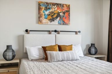 Modern neutral bedroom with upholstered headboard, textured beige bedspread, mustard velvet throw pillows and lumbar pillow, black wire-frame bedside lamps on wooden nightstands, and colorful guitar-themed artwork above the bed.