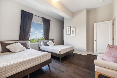 Bright modern bedroom with two twin beds in gray plaid bedding and fluffy white pillows, dark curtains framing a window with blue sky and treetop view, hardwood floors, neutral walls, framed art and a beige armchair with a mauve pillow.