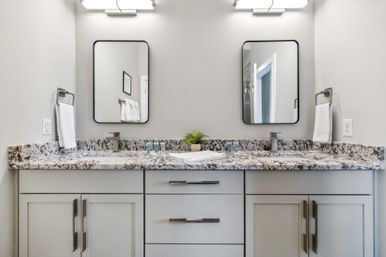 Modern double-sink bathroom vanity with gray shaker cabinets, speckled granite countertop, two rectangular mirrors, chrome faucets, wall lights, white hand towels and a small potted plant