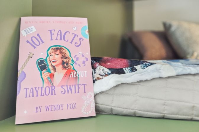 Pink '101 Facts' trivia book with illustrated smiling female singer holding a microphone on a bedside surface next to a folded blanket and pillows, cozy bedroom decor