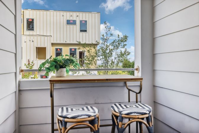 Cozy apartment balcony with a narrow wooden bar, two woven stools, potted plant and view of modern townhouses and treetops under a blue sky.