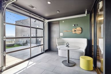 Urban rooftop lounge with glass garage door opening to a patio; white curved banquette, round table, yellow ottoman, and wooden guitar wall art on a deep green accent wall with string lights over outdoor seating.