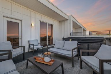 Modern rooftop patio with gray cushioned lounge chairs and sofa, coffee table with glasses and ice bucket, hot tub, and warm sunset sky