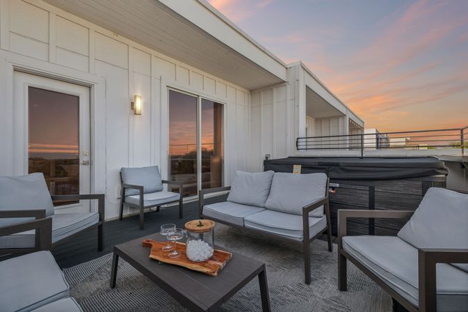 Modern rooftop patio with gray cushioned lounge chairs and sofa, coffee table with glasses and ice bucket, hot tub, and warm sunset sky