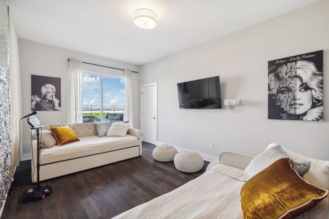 Sunlit modern apartment living room with two cream daybeds, mustard velvet pillows, wall-mounted TV, black-and-white portrait art, knitted pouf ottomans, dark hardwood floors and a glass door opening to a balcony with blue-sky view.