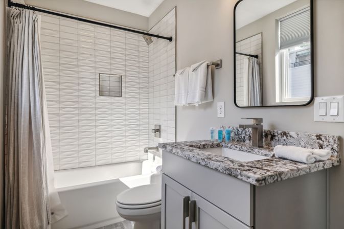 Bright modern residential bathroom with textured white-tile tub-shower, marble-look granite vanity, undermount sink, mirror, towels and window light