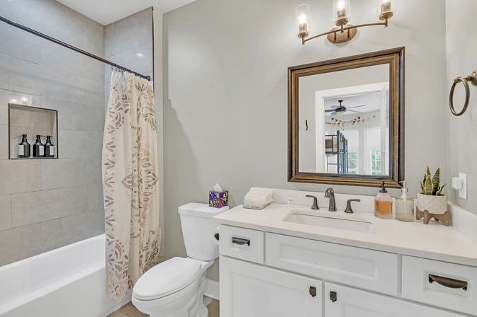 Bright contemporary bathroom — white vanity with sink and framed mirror, bathtub with patterned shower curtain, neutral tile shower niche and small potted plant.