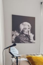 Cozy bedroom singing corner with a microphone on a stand, yellow velvet throw pillow on the bed, and a large playful black-and-white portrait of a smiling woman holding a finger to her lips on the wall.