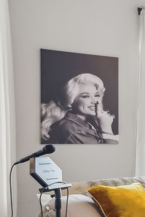 Cozy bedroom singing corner with a microphone on a stand, yellow velvet throw pillow on the bed, and a large playful black-and-white portrait of a smiling woman holding a finger to her lips on the wall.
