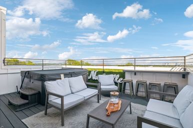Inviting rooftop deck with cushioned outdoor sofas, coffee table, hot tub and bar counter with stools overlooking a distant city skyline and blue sky with fluffy clouds.
