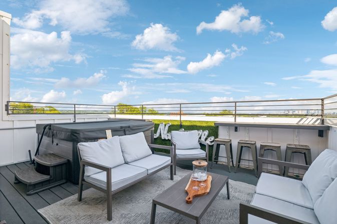 Inviting rooftop deck with cushioned outdoor sofas, coffee table, hot tub and bar counter with stools overlooking a distant city skyline and blue sky with fluffy clouds.