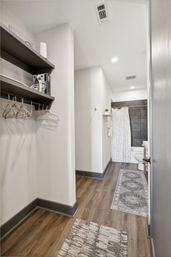 Modern apartment walk-in closet and bathroom corridor with wood-look flooring, open shelving and hangers, gray baseboards, patterned runner rugs, and a gray-tiled shower with white patterned curtain and toilet.