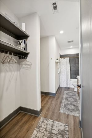 Modern apartment walk-in closet and bathroom corridor with wood-look flooring, open shelving and hangers, gray baseboards, patterned runner rugs, and a gray-tiled shower with white patterned curtain and toilet.