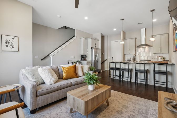 Cozy open-concept living room and kitchen with a gray sofa and fluffy pillows, mustard accent cushion, wooden coffee table with potted plant, breakfast bar with four black stools, stainless steel appliances and dark hardwood floors.